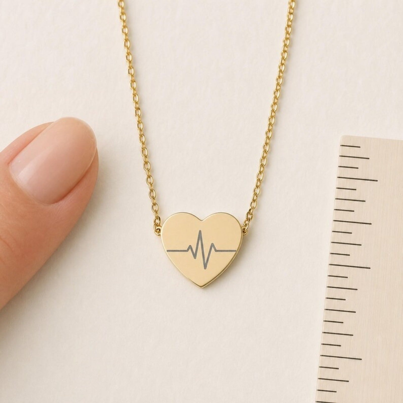 Heartbeat necklace scale reference — 18K Gold PVD heart pendant next to finger and ruler showing 0.63 in by 0.55 in. Memory That Lasts.