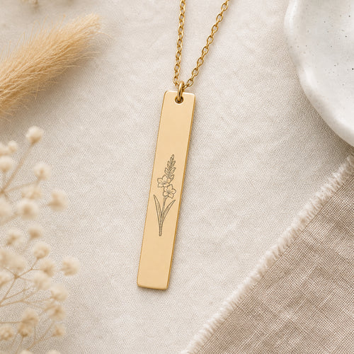 August birth flower necklace — Gladiolus engraved vertical bar pendant flatlay with dried botanicals on cream linen