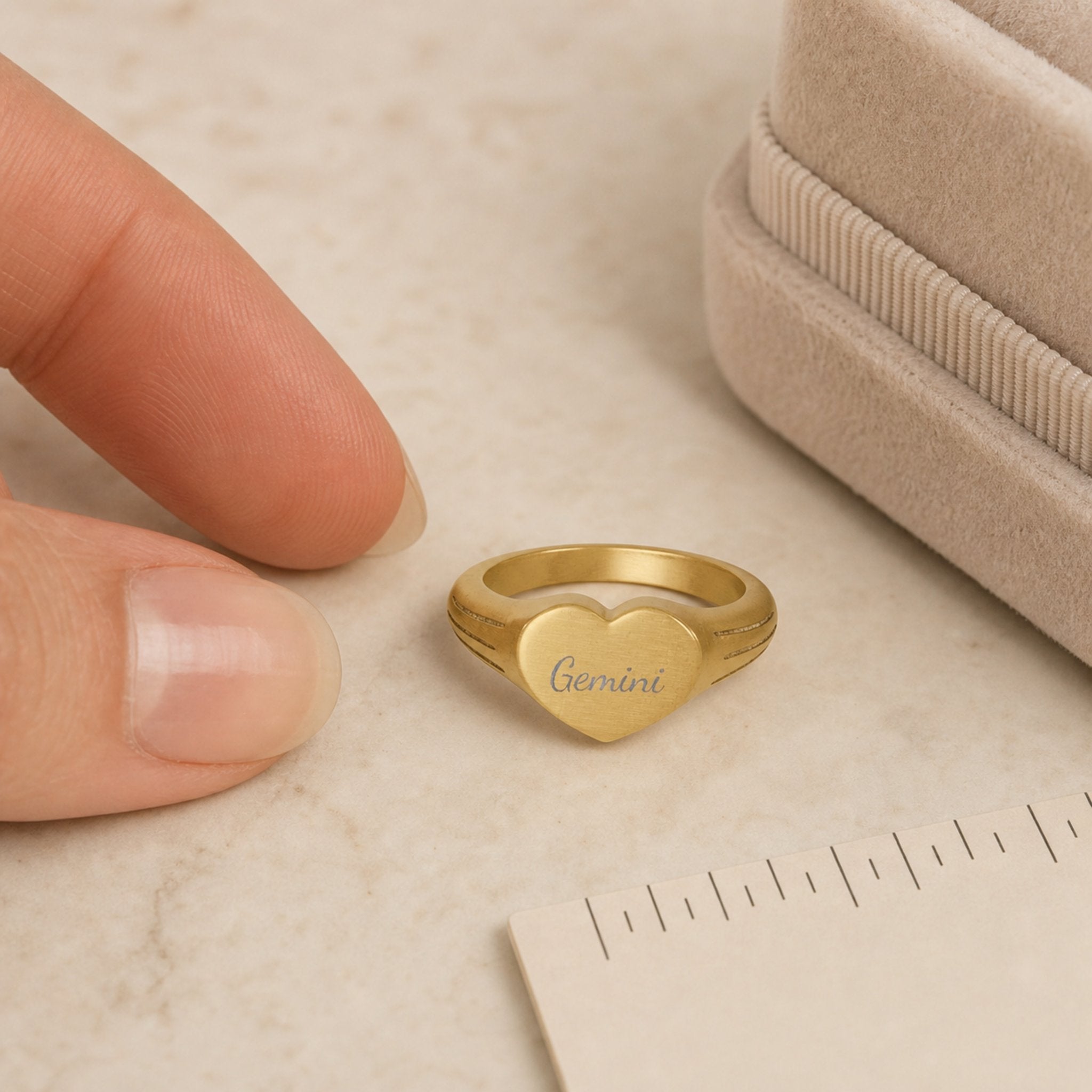 Gemini zodiac heart signet ring scale-in-hand — fingers holding heart ring next to gift box and ruler, dainty signet sizing