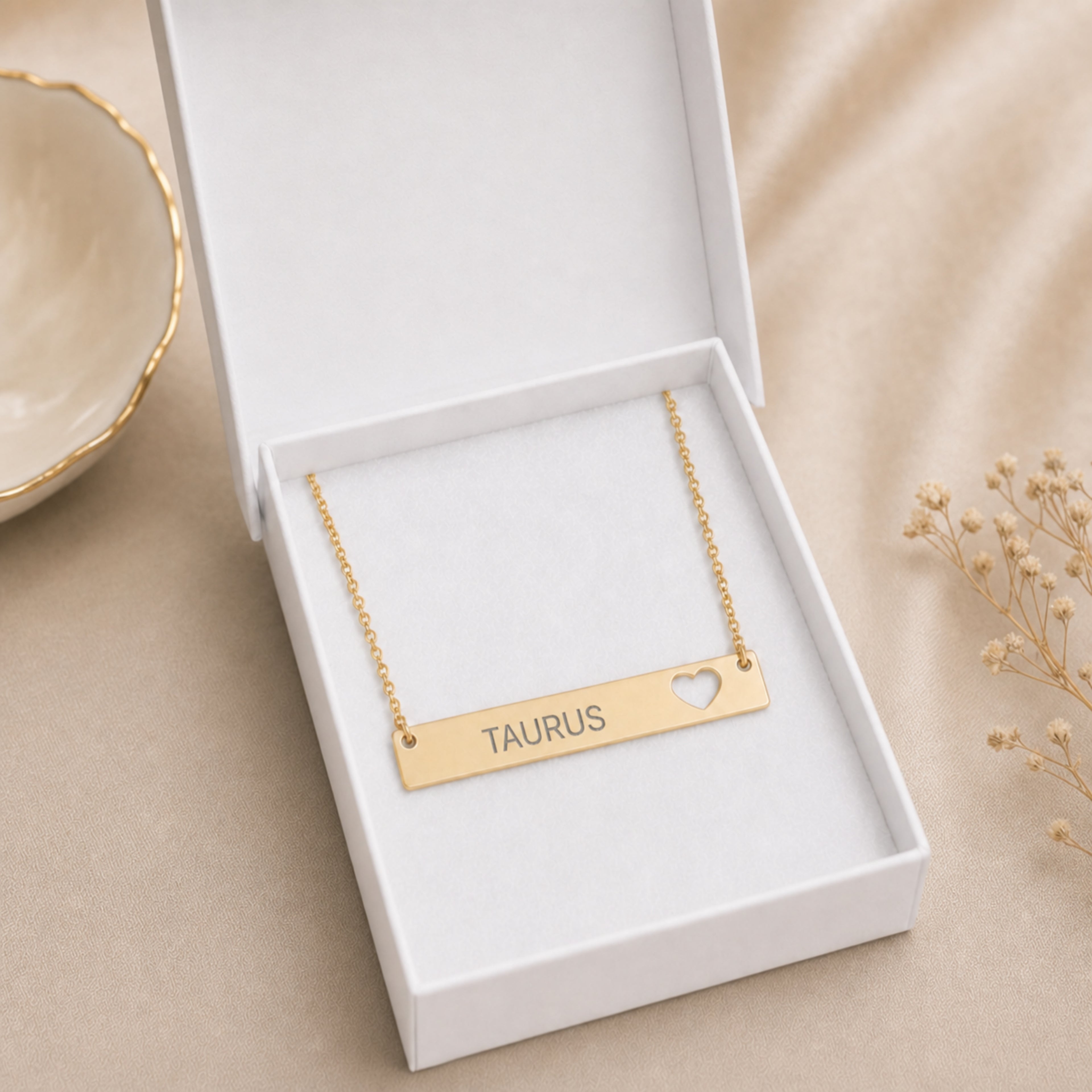 The Taurus zodiac necklace nestled in Memory That Lasts signature cream gift box — 18K Gold cutout heart bar pendant with grey laser-etched TAURUS, premium unboxing moment (AI-generated image)