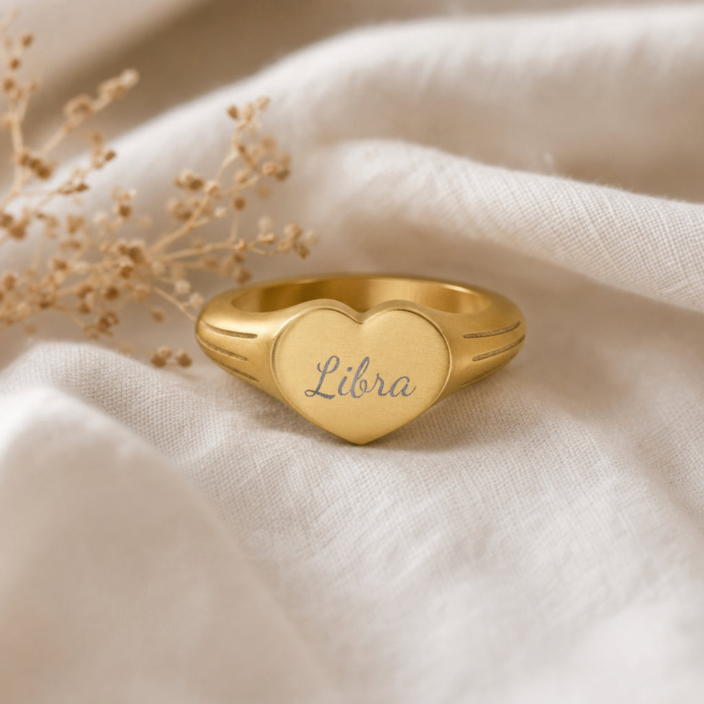Libra zodiac heart signet ring linen flatlay — engraved Libra heart ring on rumpled cream linen with dried floral, editorial styling