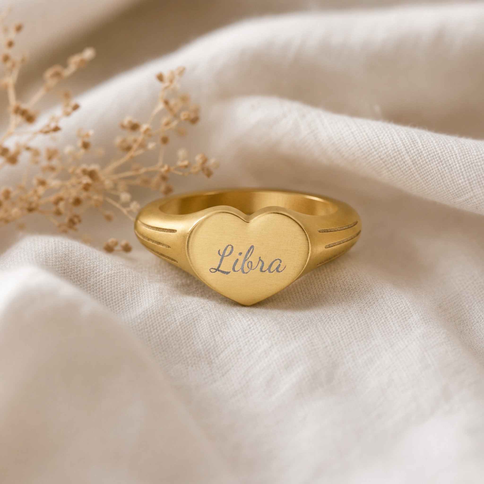 Libra zodiac heart signet ring linen flatlay — engraved Libra heart ring on rumpled cream linen with dried floral, editorial styling