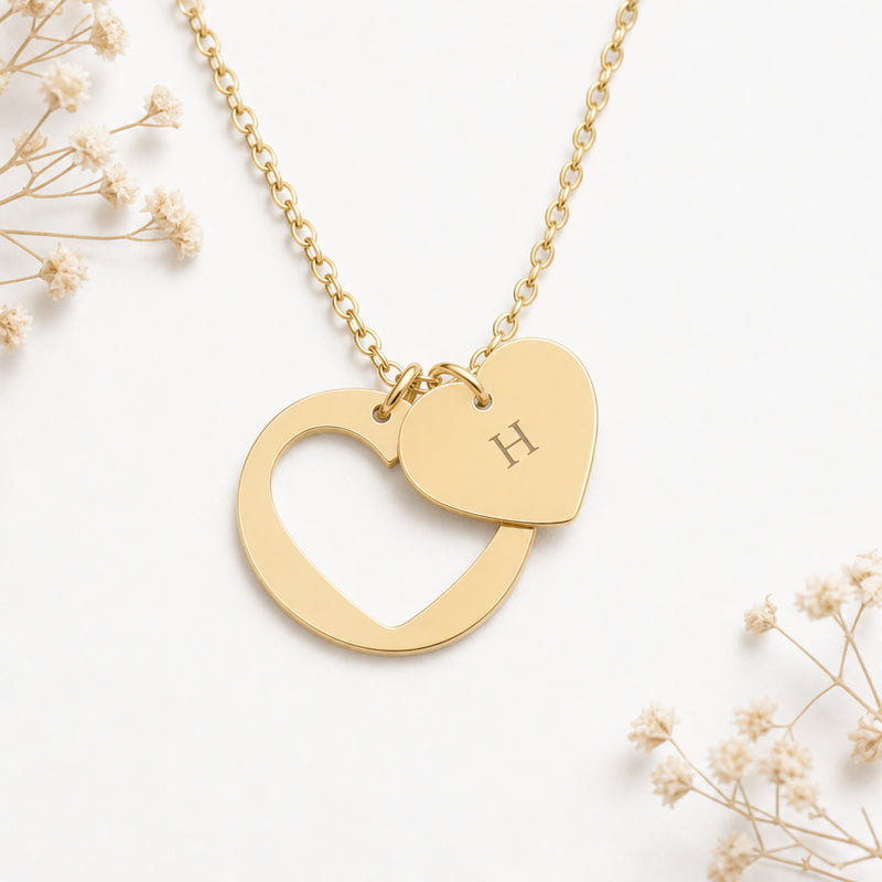 Personalized engraved initial heart necklace in 18K gold, an open heart beside a small initial charm, flat lay on linen