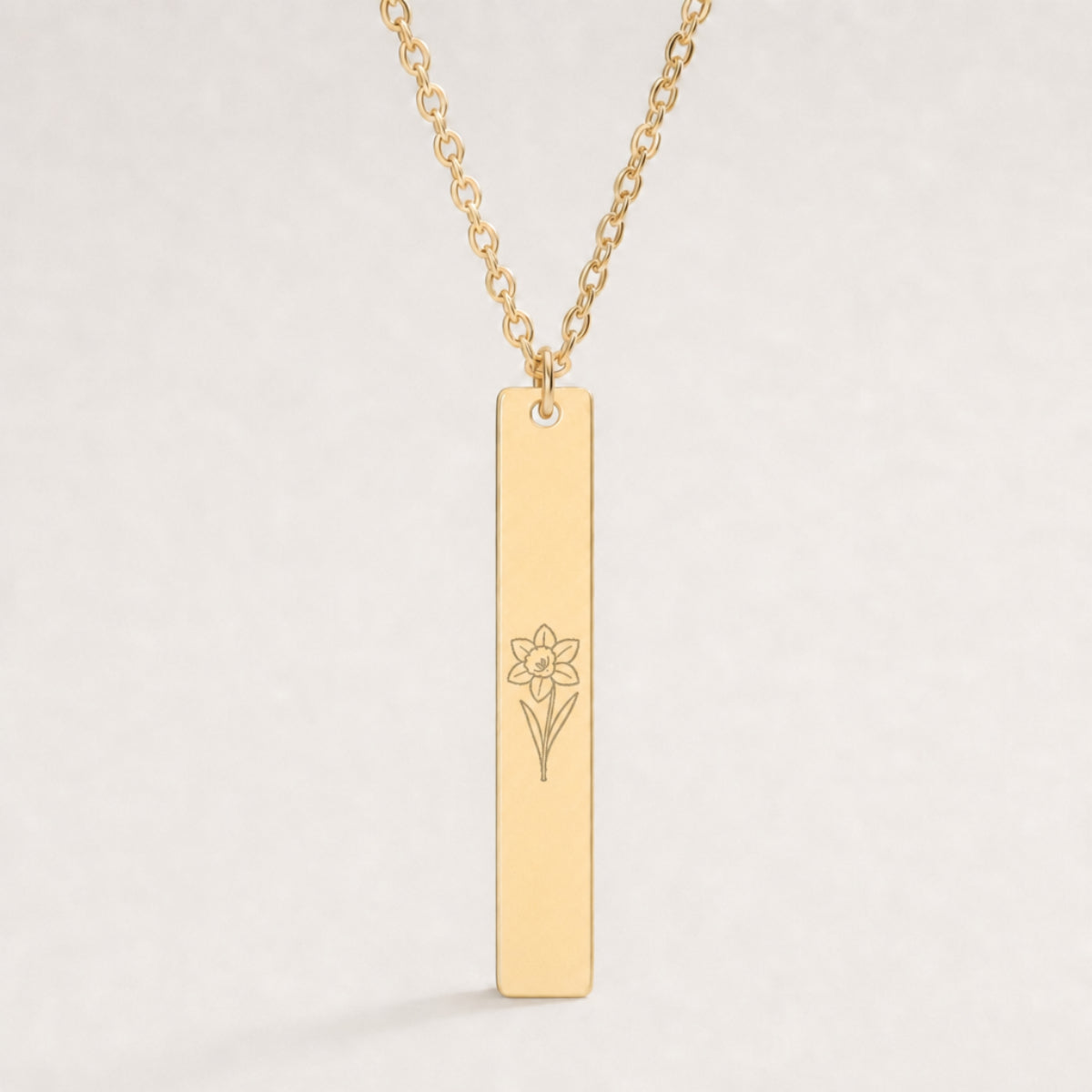 March birth flower necklace — Daffodil engraved on 1.26" × 0.20" vertical bar pendant, premium close-up macro hero, fine line art laser-etched silhouette in grey/etched matte finish on 18K Gold PVD polished mirror, premium 316L stainless steel