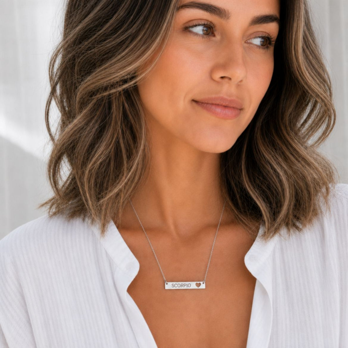Model wearing the Scorpio zodiac necklace in 316L stainless steel — polished mirror cutout heart bar with grey laser-etched SCORPIO, see-through heart cutout, white blouse, soft natural daylight