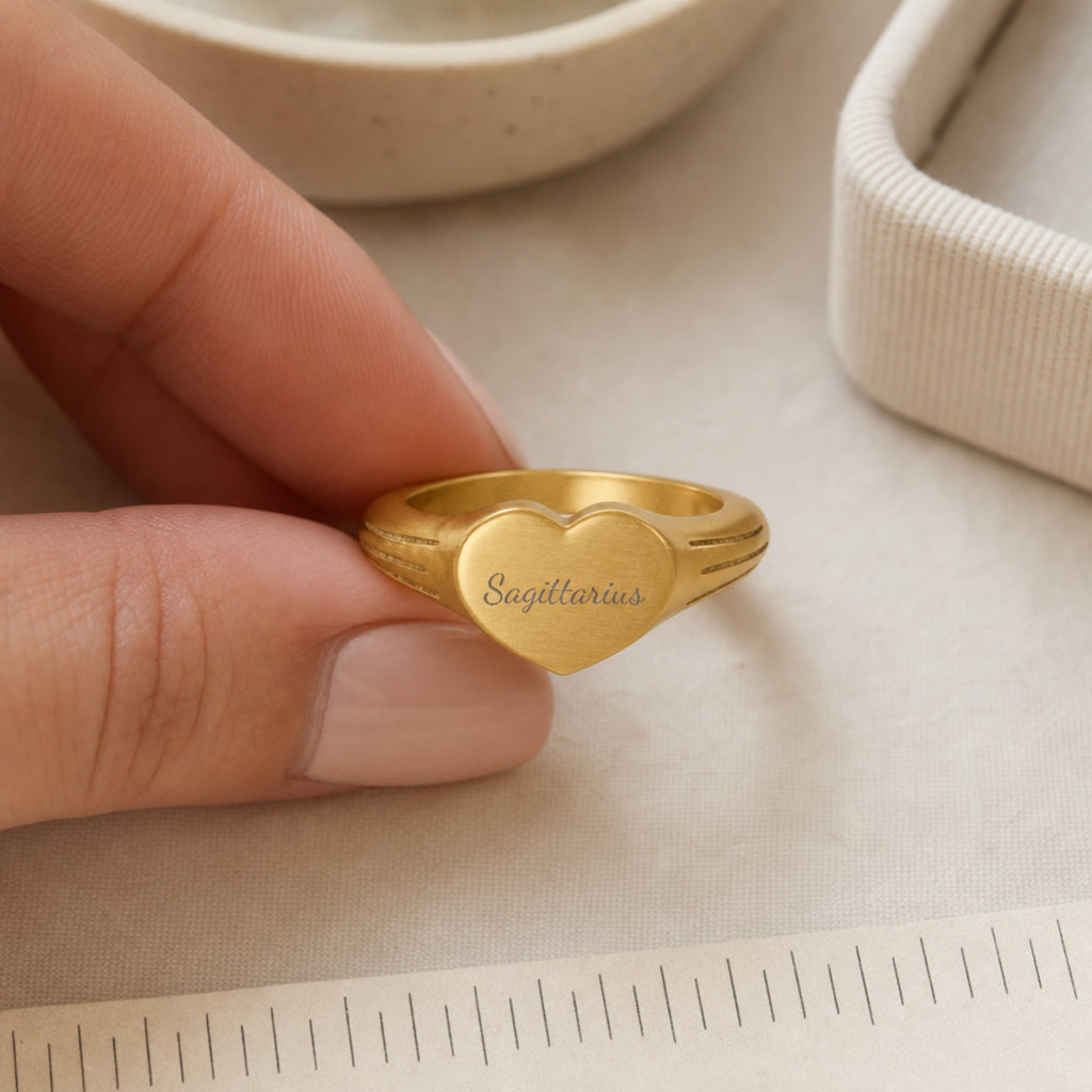 Sagittarius zodiac heart signet ring scale-in-hand — fingers holding heart ring next to gift box and ruler, dainty signet sizing