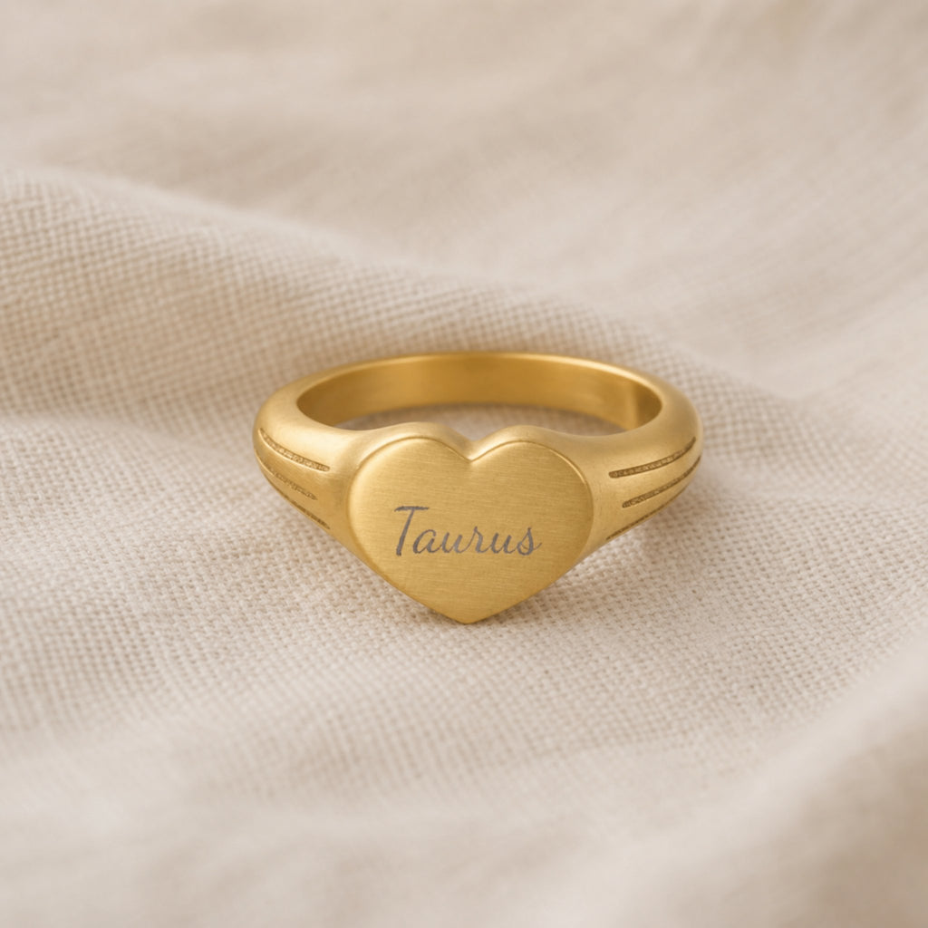 Taurus zodiac heart signet ring linen flatlay — engraved Taurus heart ring on rumpled cream linen with dried floral, editorial styling