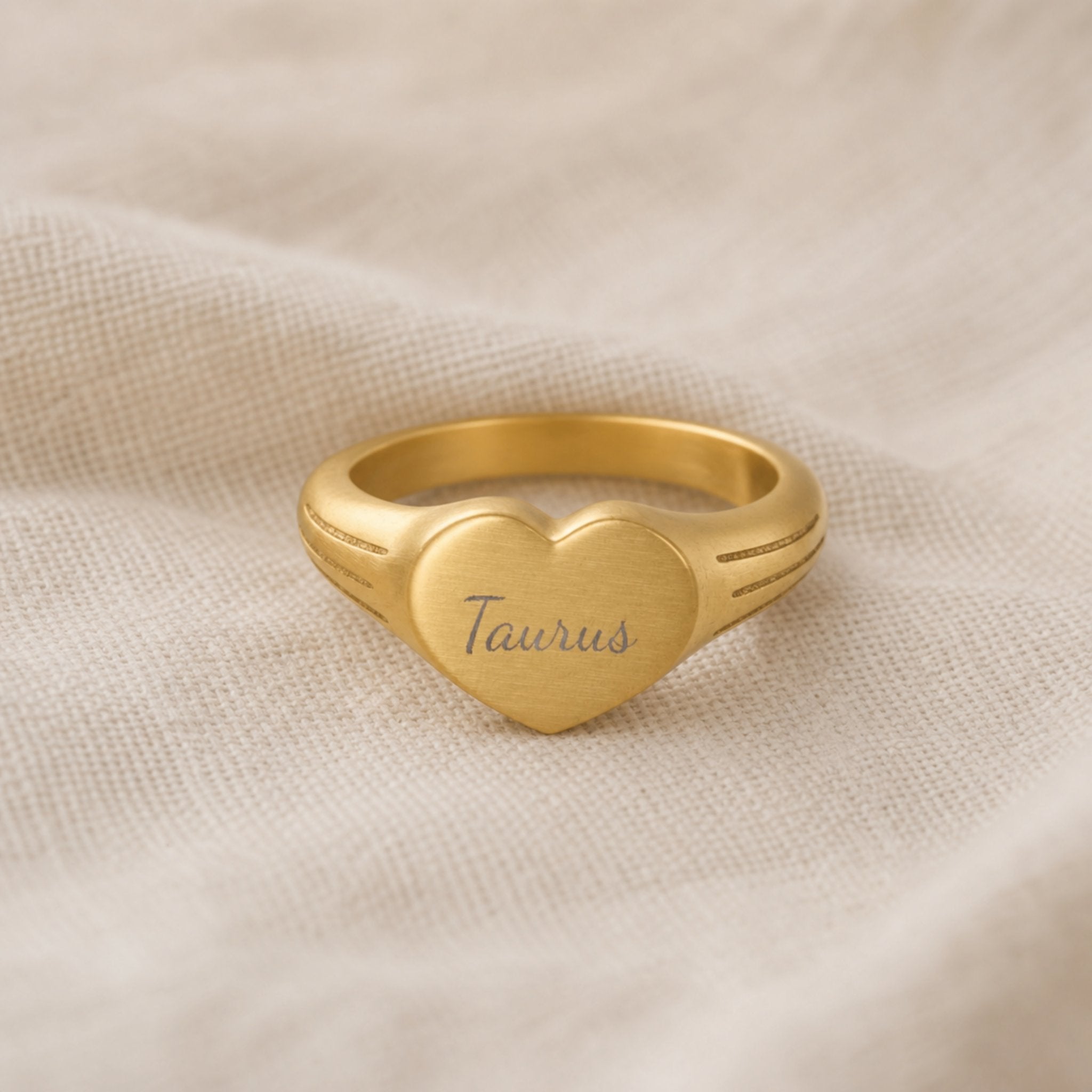 Taurus zodiac heart signet ring linen flatlay — engraved Taurus heart ring on rumpled cream linen with dried floral, editorial styling