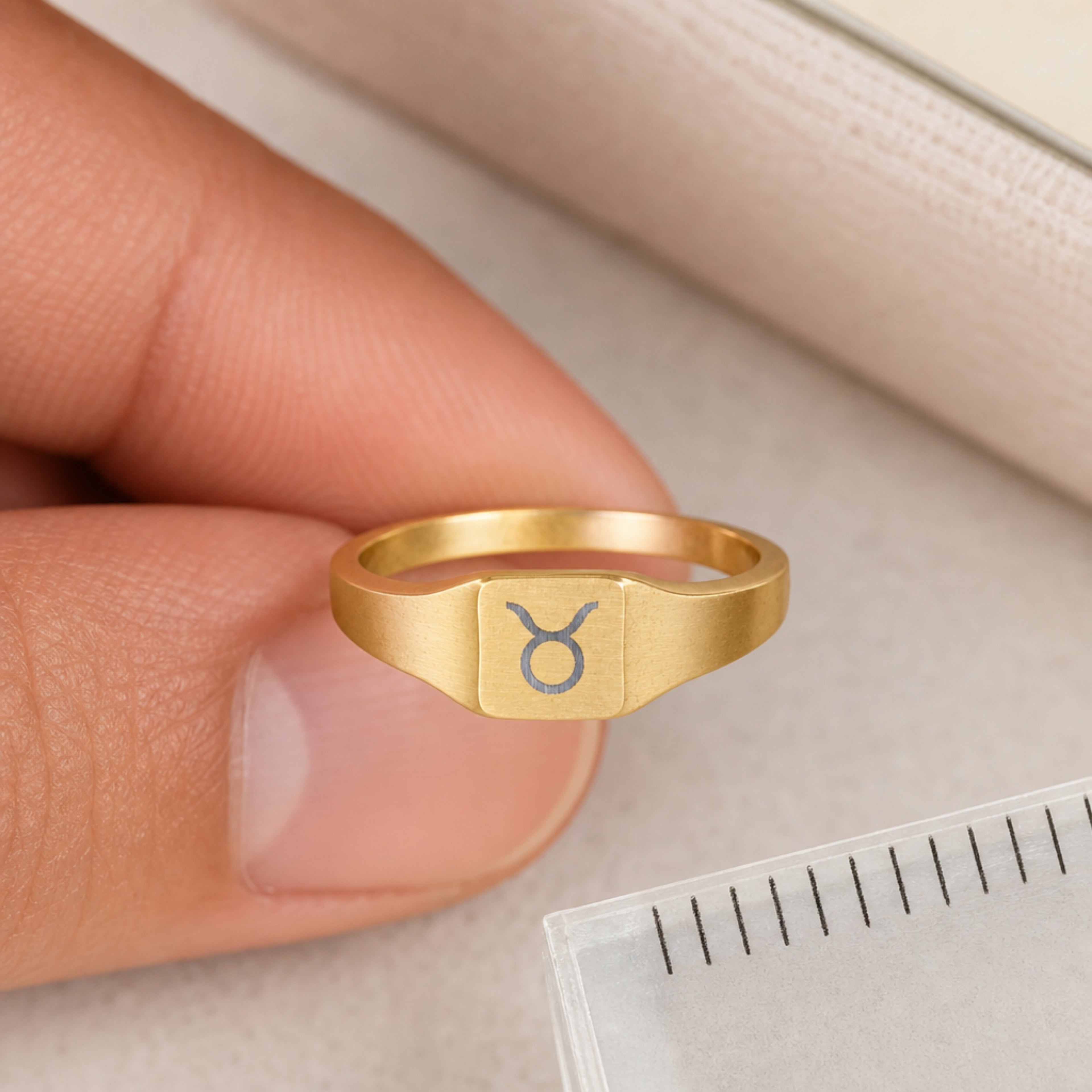 Taurus zodiac square signet ring scale-in-hand — fingers holding ring next to gift box and ruler, dainty 20mm signet sizing