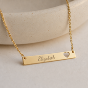 Gold personalized last name necklace, horizontal bar engraved with a name beside a cutout heart. Memory That Lasts.